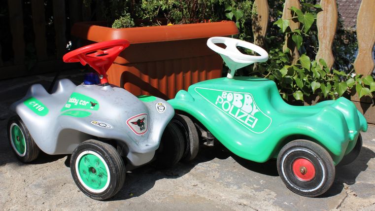 Zwei Kinderfahrzeuge stehen nebeneinander auf einem gepflasterten Boden. Das linke Fahrzeug ist silber mit roten Details, das rechte ist grün mit dem Aufschrift „Bobby Polizei“.