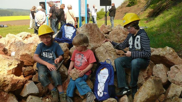 Drei Kinder mit gelben Helmen sitzen auf Felsen. Sie halten Werkzeuge und untersuchen Steine. Im Hintergrund stehen Erwachsene vor Informationstafeln.
