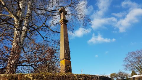 Ein hoher, grauer Obelisk steht unter einem blauen Himmel mit einigen Wolken. Um ihn herum sind Bäume und Sträucher zu sehen.