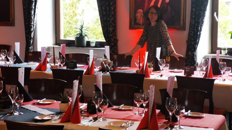 De afbeelding toont een elegant ingerichte eethoek, die waarschijnlijk is voorbereid voor een formele gelegenheid of viering. De focus ligt op verschillende lange tafels, bedekt met witte tafelkleden en rode lopers. Elke plaats heeft gepolijst bestek – vorken rechts van de borden, messen erachter en lepels helemaal links. Rode servetten, die tot driehoekige kegels zijn gevouwen, dienen als middelpunt op elk bord.