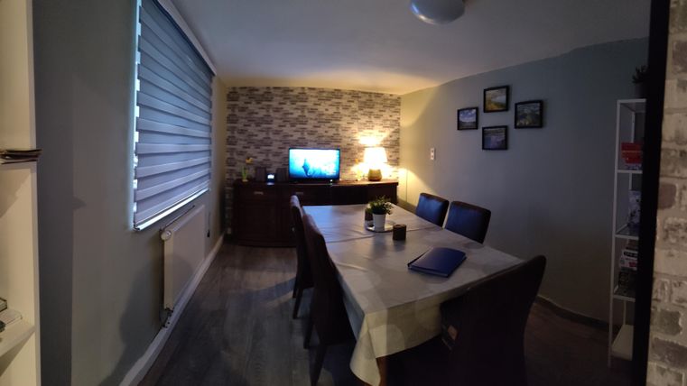 A cozy dining area with a large table and chairs. There is a television on the wall and there is a warm light source.