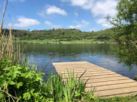 Ein ruhiger See mit einer Holzplattform im Vordergrund. Die Umgebung ist grün und die Sonne scheint durch die Wolken.