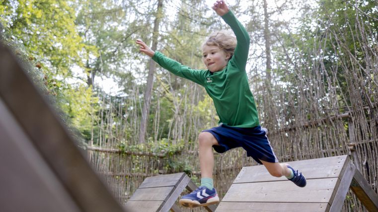 Een jongen springt vrolijk van een speeltoestel. Op de achtergrond zijn bomen en een natuurlijke speelplaats te zien.