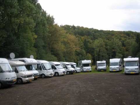 Een reeks campers staat op een parkeerplaats, omringd door bomen. De lucht is bewolkt en het landschap lijkt rustig en idyllisch.