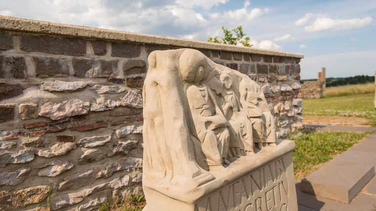 Eine Sandsteinskulptur mit Figuren, die in einer Szene vereint sind, steht vor einer Mauer aus Steinen. Der Himmel ist bewölkt und es ist ein grünes Feld im Hintergrund sichtbar.