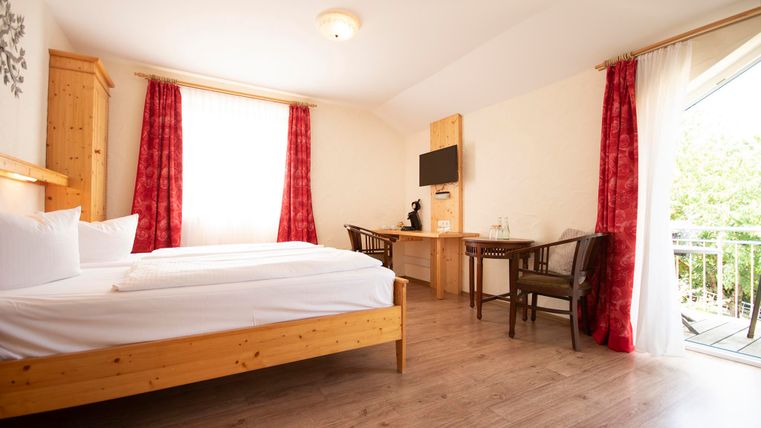 A bright room with a double bed, a table, and chairs. Large windows provide plenty of daylight and a view outside.