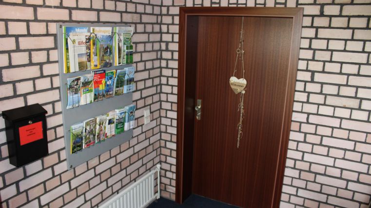 Ein kleiner Eingangsbereich mit einer Ziegelmauer. An der Wand hängen Flyer und eine Tür mit einem herzförmigen Anhänger.