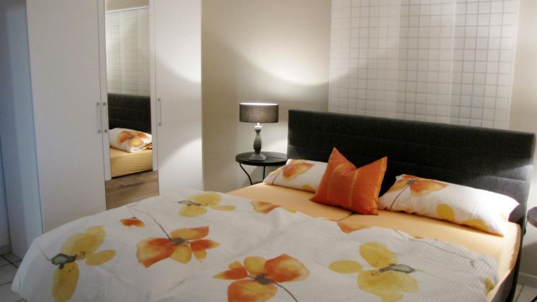 A modern bedroom with a large double bed and colorful floral decorations. In the background, there is a mirror and a lamp on a bedside table.
