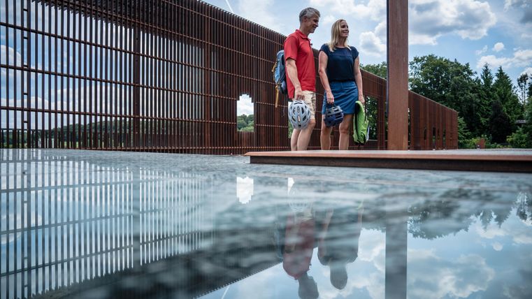 A couple stands on a platform with reflective water. In the background, there are trees and a blue sky with clouds.