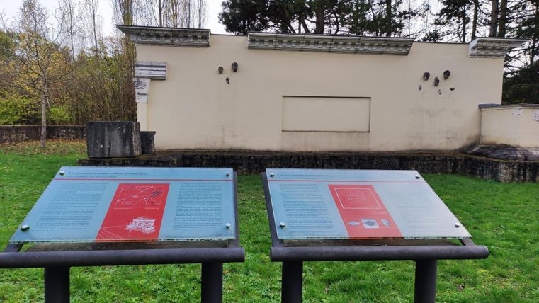 Two explanatory panels stand in front of an old wall in a park-like setting. In the background, trees can be seen, and the area is covered with green grass.