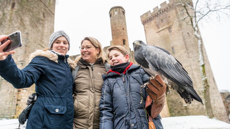 Drei Personen posieren für ein Selfie vor der Kasselburg. Ein Junge hält einen Greifvogel auf dem Arm. Es liegt Schnee auf dem Boden.