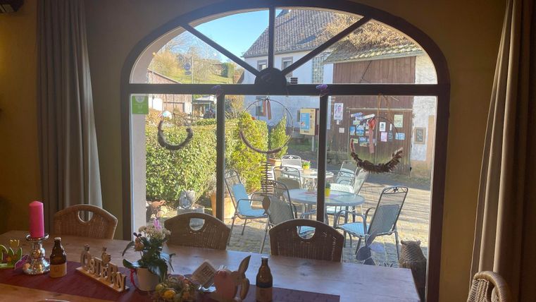 Ein einladender Innenraum mit großen Fenstern, die auf einen Außenbereich mit Tischen und Stühlen blicken. Im Vordergrund steht ein gedeckter Tisch mit Blumen und Dekoration.