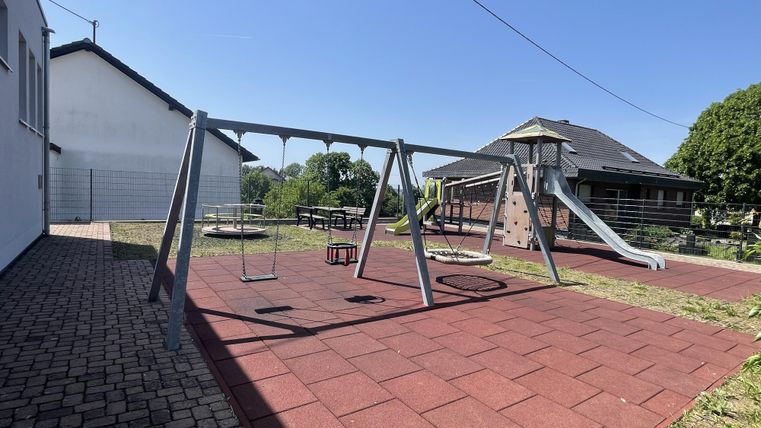 Een speelplaats met een schommel en een glijbaan. De vloer is bedekt met rode rubberen tegels en er is veel grasgebied.