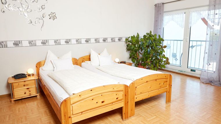 Een gezellige slaapkamer met twee houten eenpersoonsbedden. Er is een nachtkastje, een plant en grote ramen met uitzicht naar buiten.