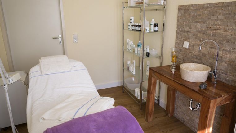 Ein ruhiger Behandlungsraum mit einem Massagebett und einer Holztheke. Im Hintergrund steht ein Regal mit Pflegeprodukten.
