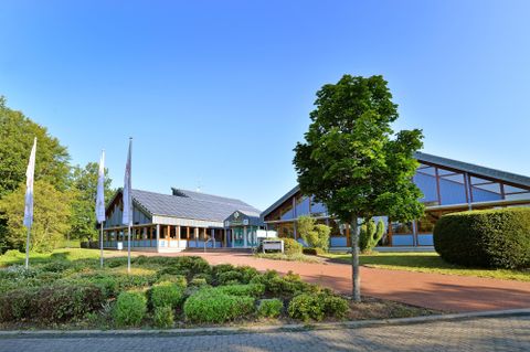 Een modern gebouw met grote ramen en een verzorgde buitenruimte. Omringd door bomen en struiken onder een heldere blauwe lucht.