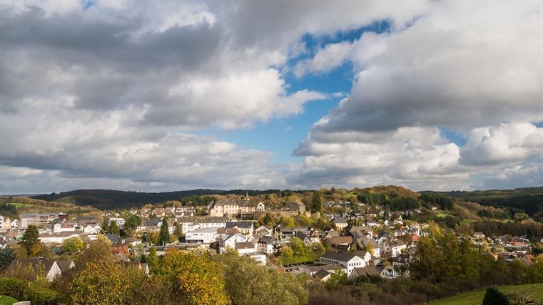 Ein malerisches Dorf umgeben von Hügeln und Bäumen. Der Himmel ist teils bewölkt, mit vereinzelten sonnigen Bereichen.