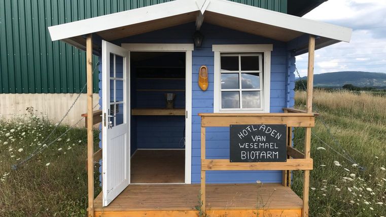 Een klein blauw verkooppaviljoen met een houten voortent staat in een open landschap. Het toont een bord met de tekst "Hotel-laden van Wesemel Biotarm".