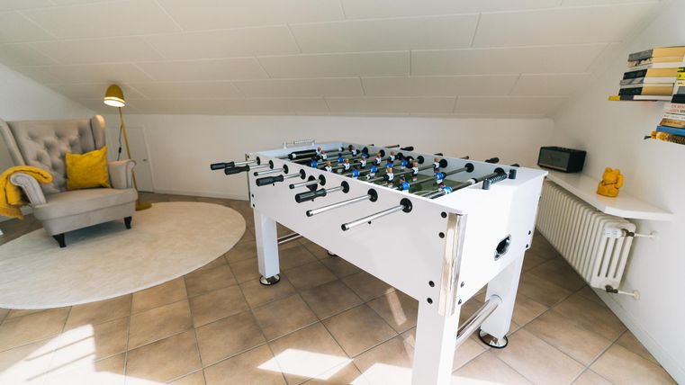 Een moderne tafelvoetbaltafel staat in een lichte ruimte met cerumnkleurige vloer. Op de achtergrond zie je een gezellige stoel en enkele boekenplanken.