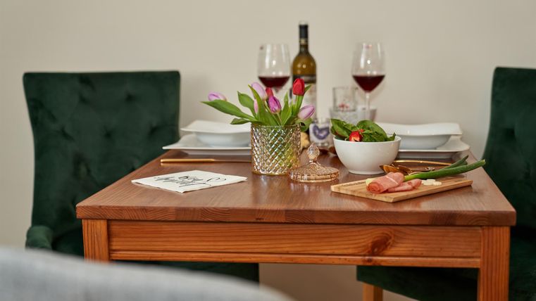 A beautifully set table with red wine and fresh ingredients. In the background, there are green chairs and a decorative vase.