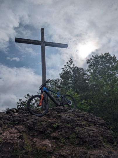 Een fiets staat op een rots voor een houten kruis. De lucht is bewolkt en er zijn bomen op de achtergrond te zien.