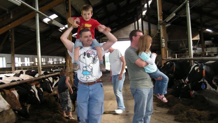 A father is carrying his child on his shoulders in a barn. In the background, cows and other people can be seen.