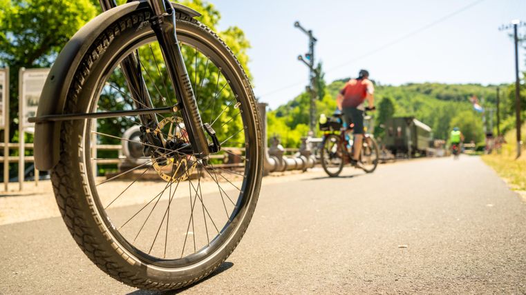 Een fietswiel staat op de voorgrond op een zonnige weg. Op de achtergrond rijden twee fietsers langs de bosrand.