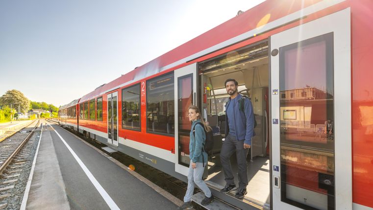 Two people are getting off a red train at a modern train station. The surroundings are bright and friendly with a clear sky.