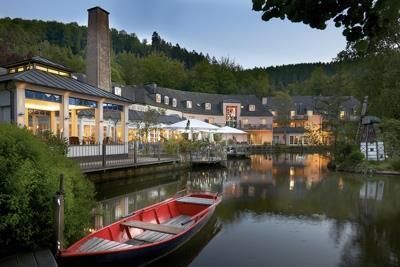 A beautiful hotel by the water with a red boat. The surroundings are green and the atmosphere is calm and inviting.