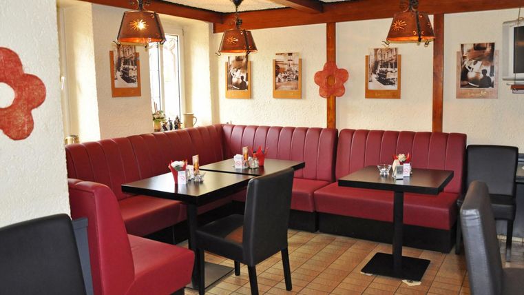 Ein gemütliches Restaurant mit roten Sitzbänken und Tischen. An den Wänden hängen Bilder und es gibt dekorative Lampen.