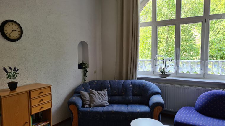Ein gemütliches Wohnzimmer mit einem blauen Sofa und einem lila Sessel. Große Fenster sorgen für viel natürliches Licht und Blick ins Grüne.