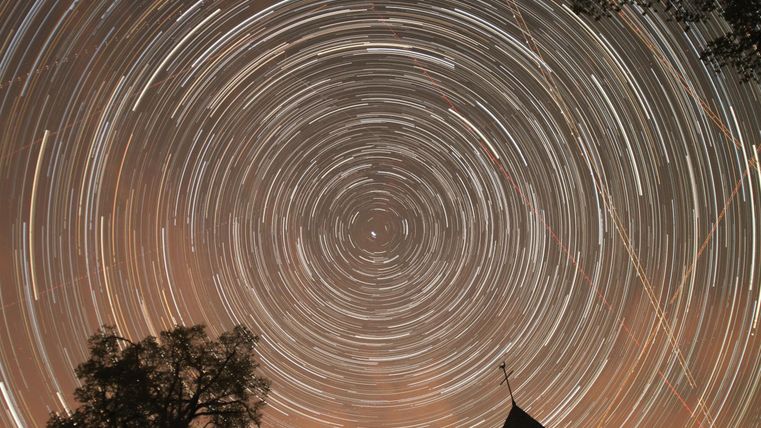 Een fascinerende sterrenhemel met spiraalvormige sterrenstrepen. Op de voorgrond zijn silhouetten van bomen en een dak zichtbaar.