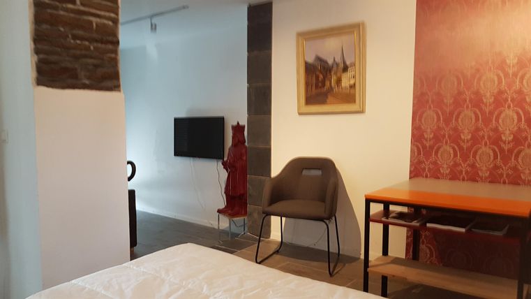 Ein modernes Zimmer mit einem Bett, einem Tisch und einem Fernseher. An der Wand hängt ein Bild und die Farben sind hell und freundlich.