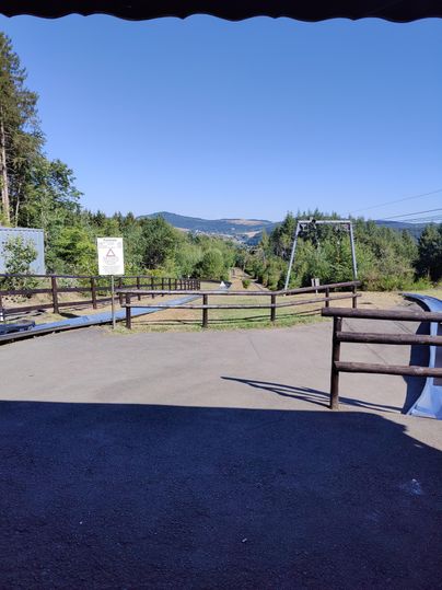 A tranquil landscape with trees and a clear blue sky. In the foreground, a fenced path leading into nature is visible.