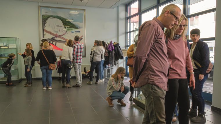 People in the Gerolsteiner Visitor Center are looking at an exhibition. Some are reading information, others are chatting. In the background, there is a large picture.