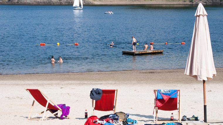 Ein schöner Strand mit Liegestühlen und einem Sonnenschirm. Im Wasser schwimmen Menschen und ein Segelboot ist in der Ferne zu sehen.