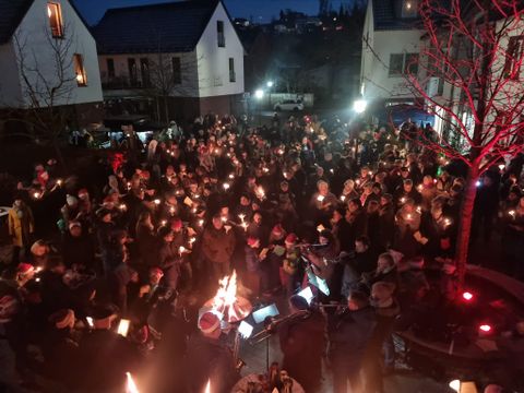 Eine große Menschenmenge versammelt sich in einem Hof, jeder mit einer brennenden Laterne. Die Stimmung ist festlich und die Umgebung ist von schwachem Licht erhellt.