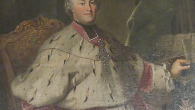 Een oudere man in traditionele kledij zit op een weelderige stoel. Hij draagt een ketting en heeft een ernstige, maar vriendelijke uitdrukking.