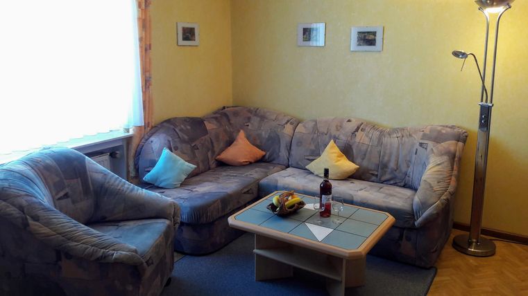 Een gezellige woonkamer met een grote bank en een comfortabele stoel. Op de tafel staan fruit en een fles wijn, terwijl het licht zachtjes door het raam naar binnen valt.