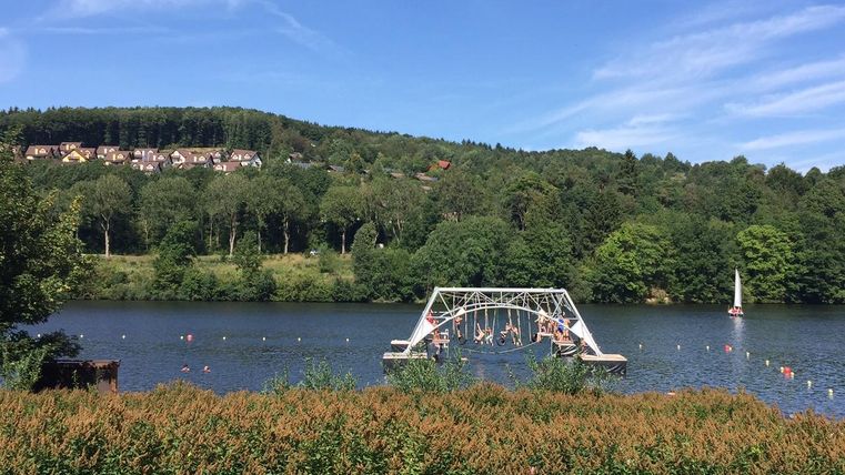 Ein ruhiger See umgeben von Bäumen und Wiesen. Im Hintergrund sind Menschen, die auf einem Sprungbrett spielen, und ein Segelboot zu sehen.