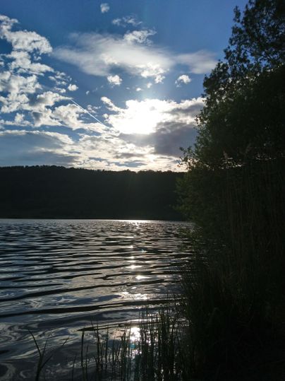 Een rustige meer bij zonsondergang, omringd door bomen en een heldere lucht. Het wateroppervlak weerspiegelt de wolken en het licht van de zon.