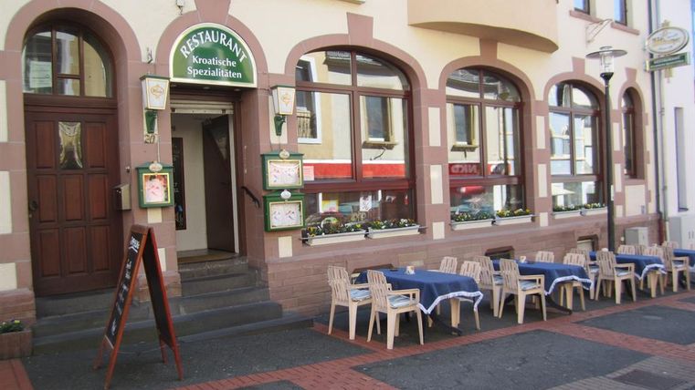 Exterior view of a restaurant offering Croatian specialties. Tables with blue tablecloths and chairs are set up on the sidewalk.