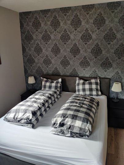 A modern bedroom with a double bed and a stylish patterned background. The bedcovers are in a checked design and next to the bed are small lamps.