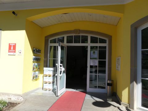 De ingang van een lichtgele gebouw met een open glazen deur. Een rode loper leidt naar binnen, en enkele informatiebrochures zijn ernaast bevestigd.