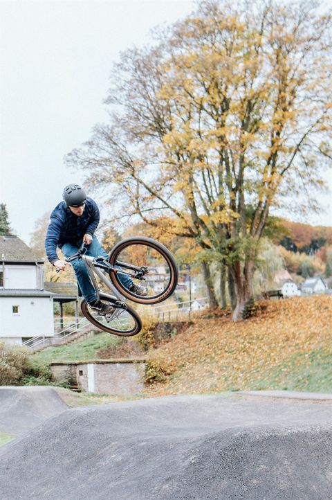 Een BMX-rijder springt over een ramp in een park. Op de achtergrond zijn bomen met herfstkleuren te zien.