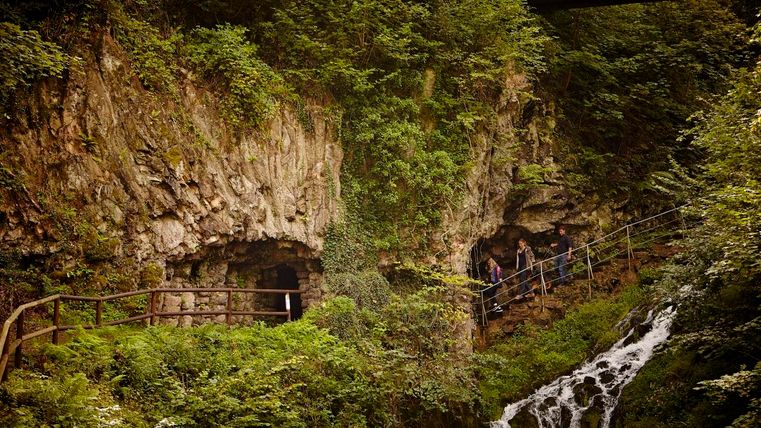 Eine malerische Landschaft mit steilen Felsen und einem kleinen Wasserfall. Wanderer erkunden den Weg entlang des Wassers.