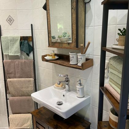 Een moderne badkamer met een witte wastafel en een houten spiegel. Op de achtergrond zijn handdoeken en decoratieve elementen op planken te zien.