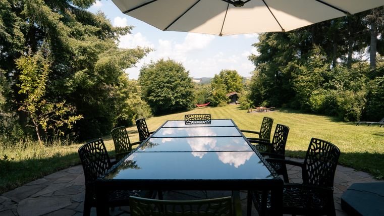 Ein eleganter Tisch mit Stühlen steht im Freien unter einem großen Sonnenschirm. Im Hintergrund sind grüne Bäume und eine weite Wiese zu sehen.