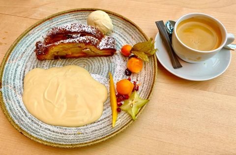 A plate with apple strudel, cream, and colorful fruit. There is also a cup of coffee in a light saucer.