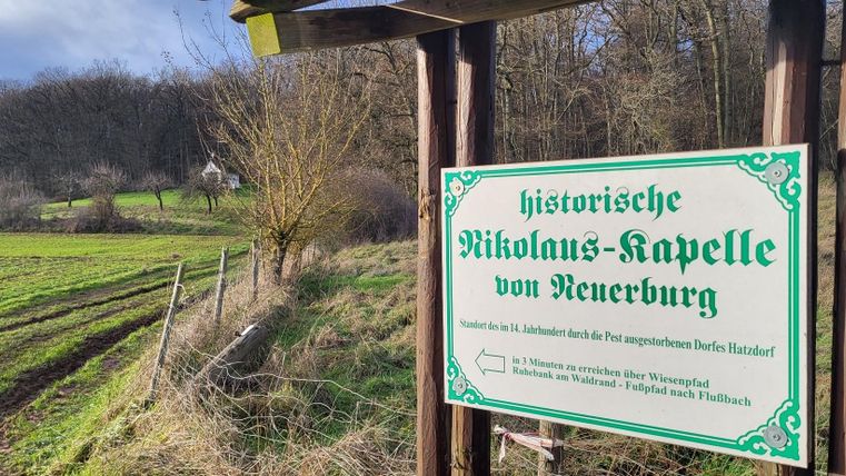 Ein Schild zeigt die historische Nikolaus-Kapelle von Wenerburg. Im Hintergrund sind grüne Felder und Bäume zu sehen.